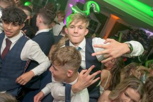 2 students perform a piggy-back whilst celebrating at their school prom. Mobile disco lights fill the background. A DJ plays music and the crowd are dancing.