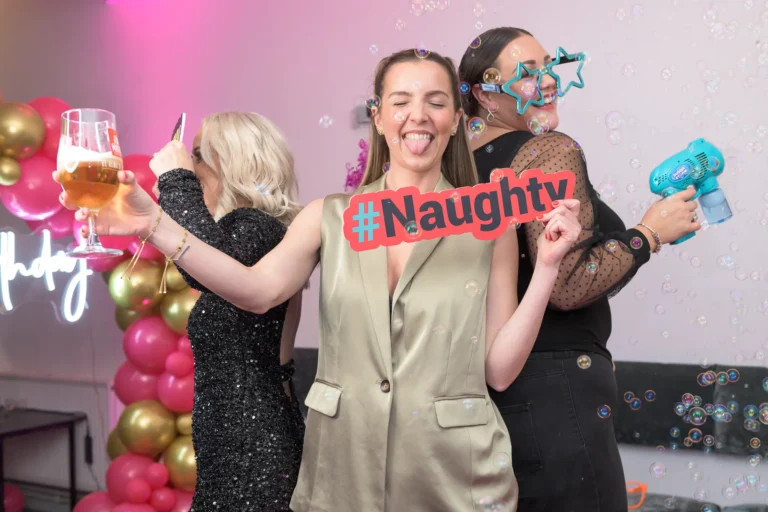 A lively group of friends celebrate on the 360 Photo Booth, capturing a fun birthday video as the camera spins around them. They're laughing, firing bubble guns, and showing off silly props like party hats and giant sunglasses — the perfect mix of chaos and celebration.