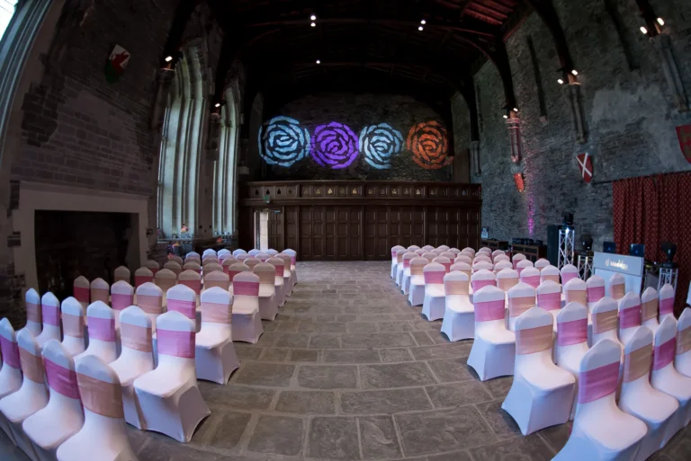 Wedding aisle in castle setting. Chairs decorated with pink covers. Disco lights project roses on the wall. Wedding DJ setup ready to play music for the procession.