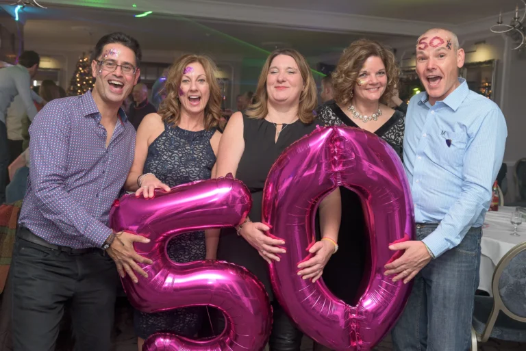 Friends and family at 50th birthday party. They are holding balloons and smiling. Crowd are dancing to music in the background.