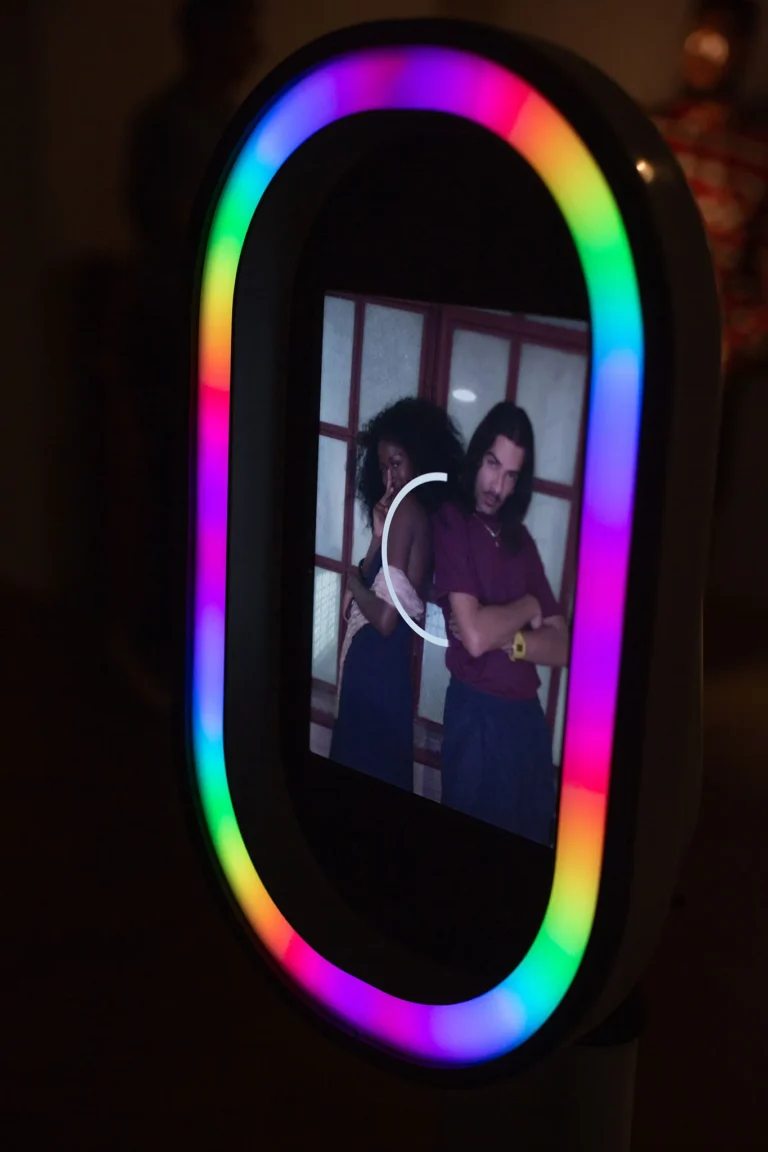 Salsa photo booth pod displays rainbow pattern. 2 people are seen taking a selfie in the reflection.
